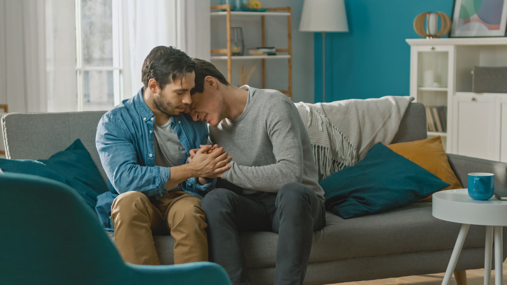 Male couple sitting together on a couch and grieving. One has his head on the others shoulder and is crying.