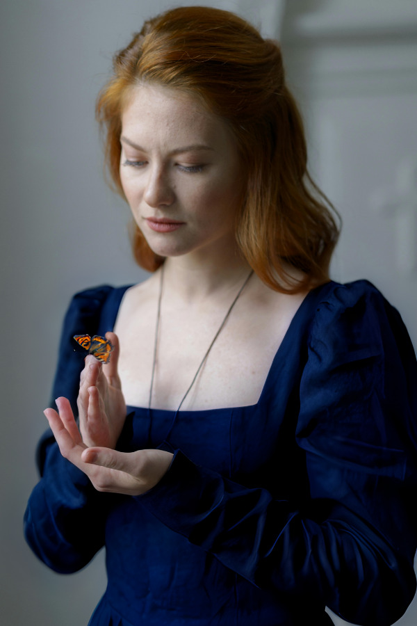 Melancholic red-haired woman wearing a blue dress and looking at a monarch butterfly on her hands in front of her.