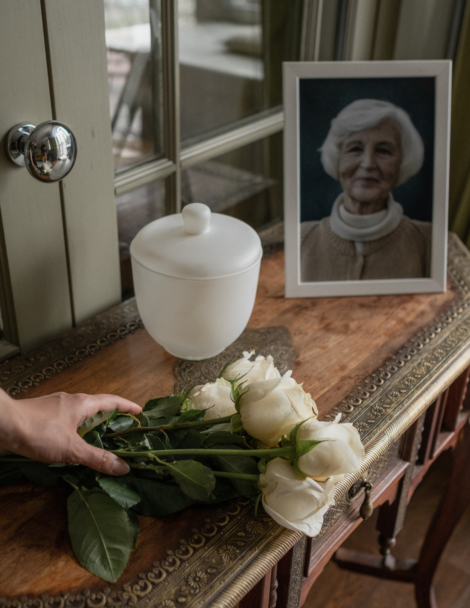 A hand sets a bouquet of white roses on a wooden table next to a framed picture of an elderly woman and a white urn.