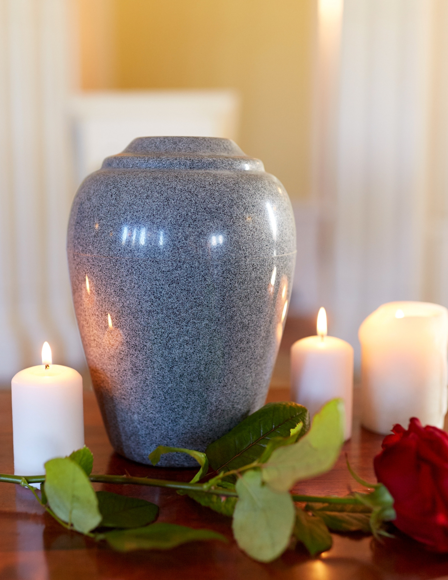 A stone urn on a wood table surrounded by three lit candles and a rose.