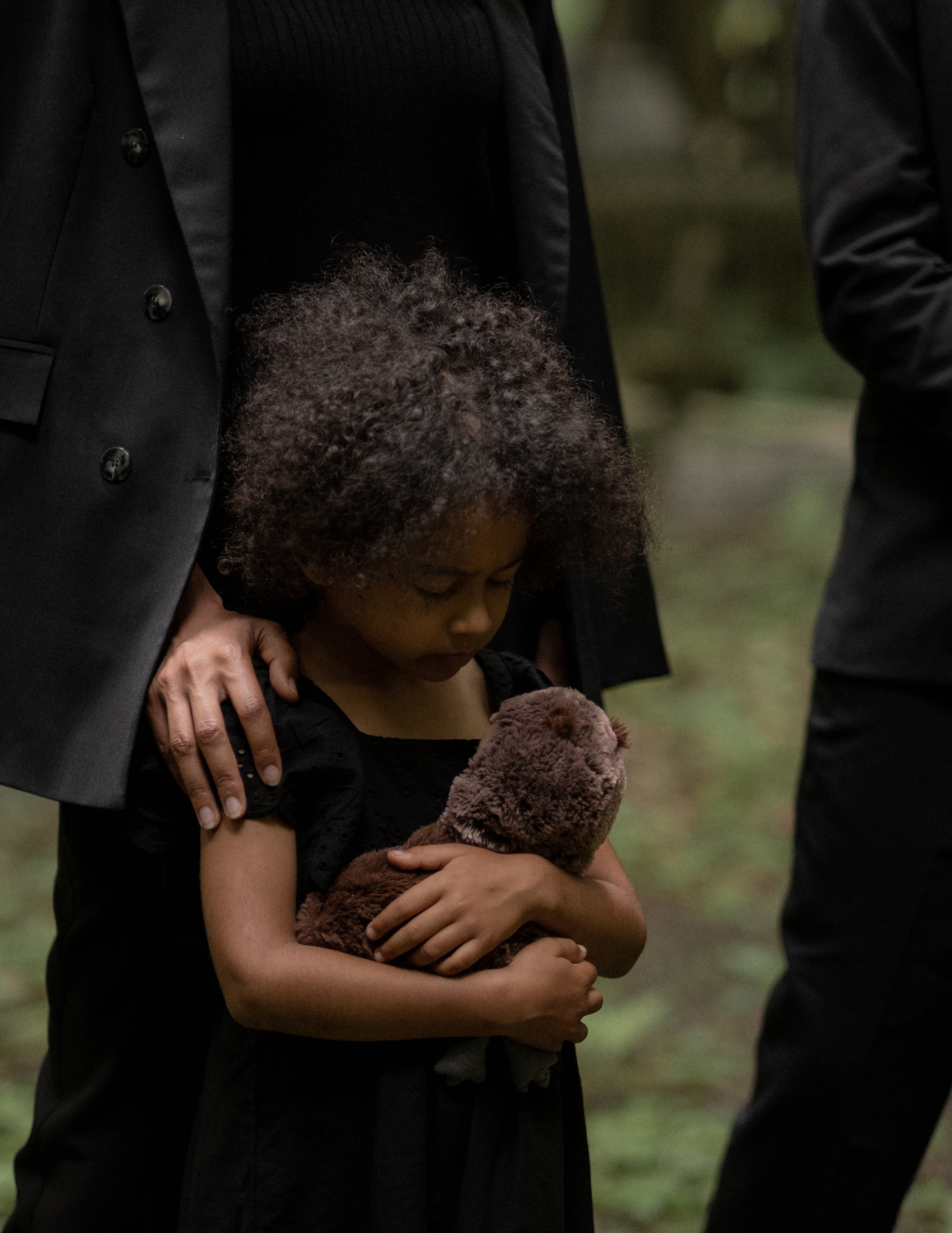 A young girl in black holds a stuffed animal while an adult rests a hand on her shoulder.