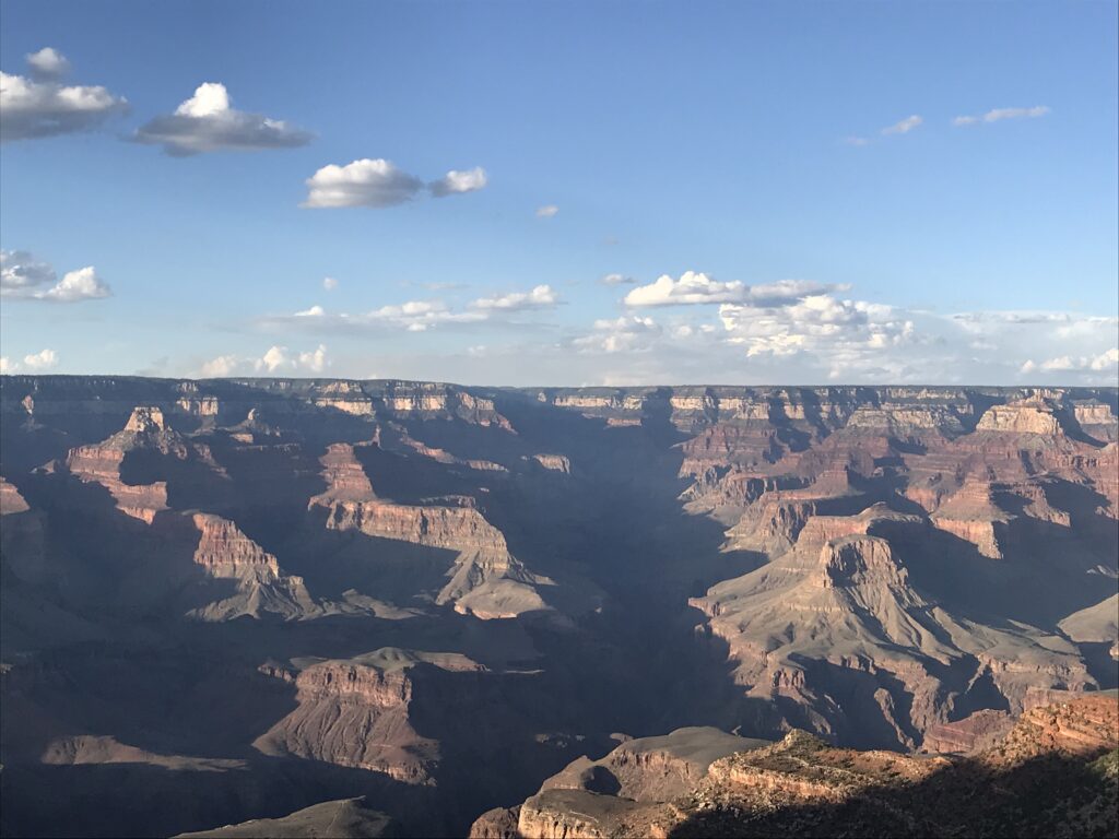 A wide desert canyon landscape with layered cliffs, deep shadows, and a bright blue sky.