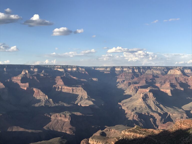 A wide desert canyon landscape with layered cliffs, deep shadows, and a bright blue sky.