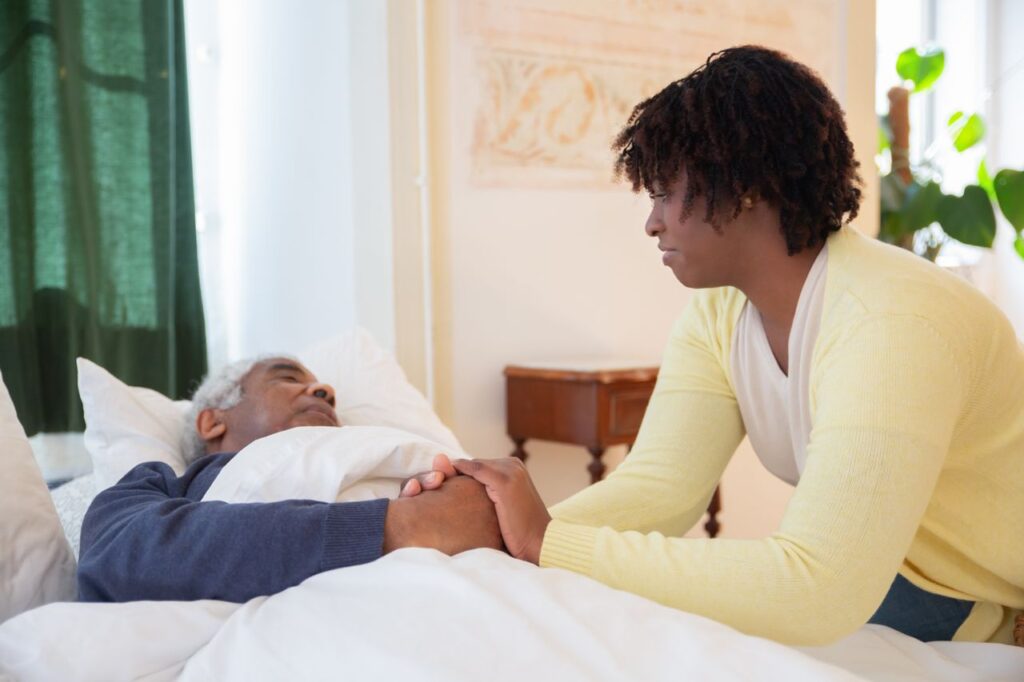 A woman gently holds the hand of an older man resting in bed