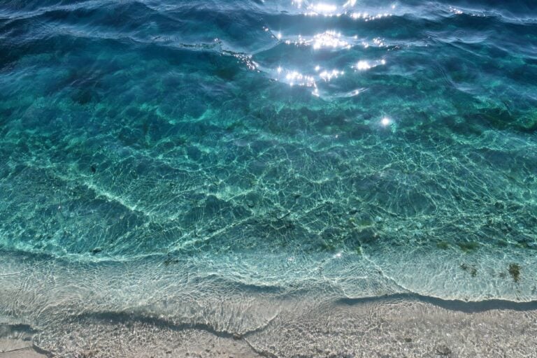 Clear turquoise ocean water gently meeting a sandy shore in bright sunlight.