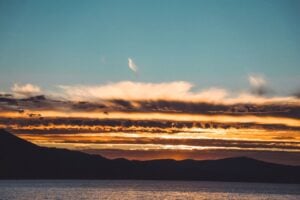 A golden sunset over distant mountains and calm water beneath a blue sky.