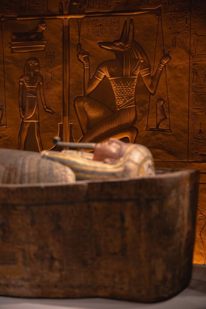 A dimly lit Egyptian display featuring Anubis imagery, hieroglyphs, and a sarcophagus in the foreground.
