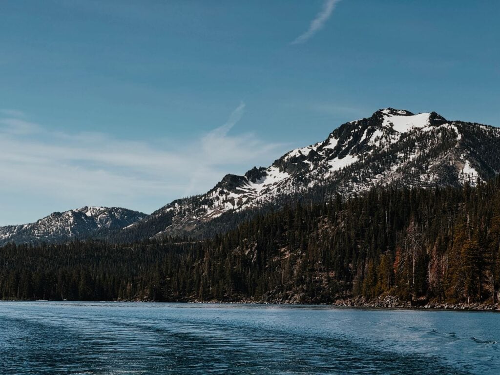 A clear mountain lake bordered by dense forest and snow-dusted peaks under a blue sky.