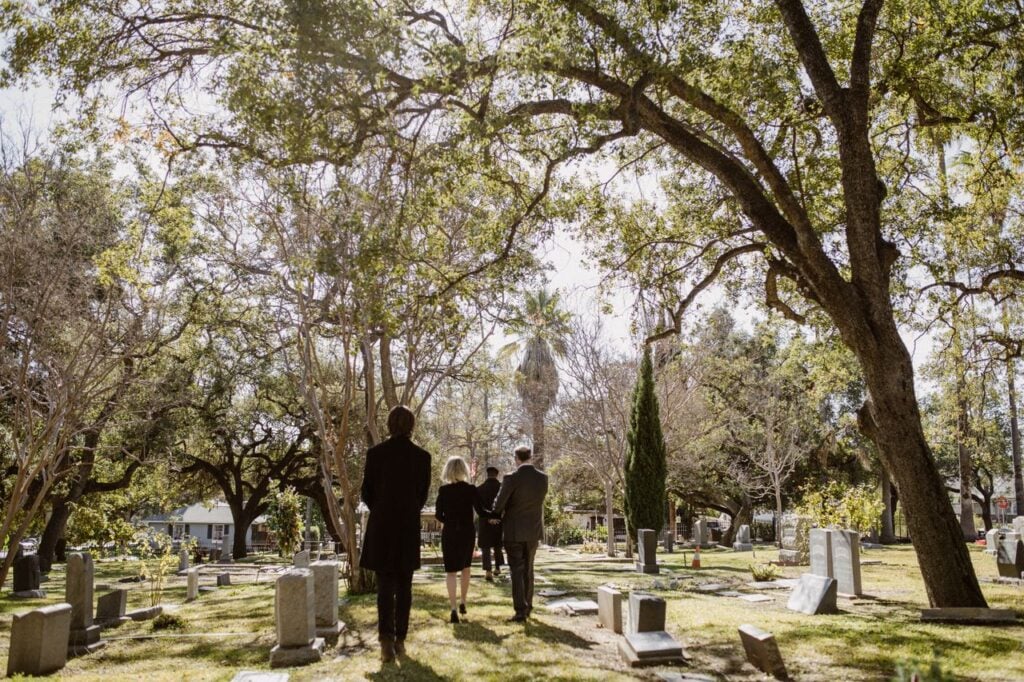 A small group walking through a peaceful cemetery surrounded by trees and sunlight.