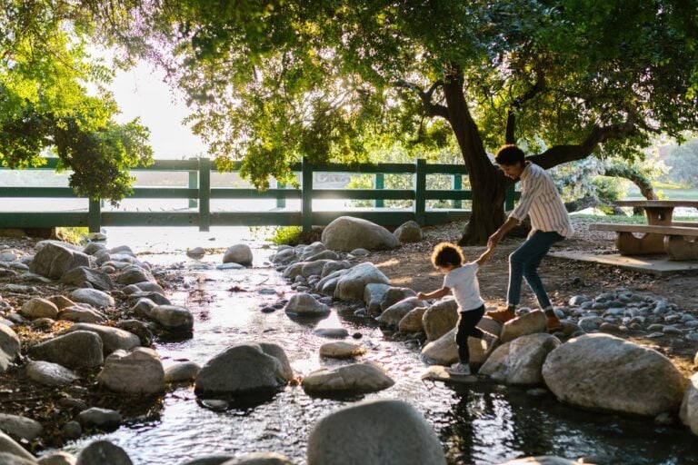 A parent and child cross a shallow creek surrounded by rocks and sunlit trees.