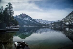 A calm mountain lake surrounded by pine trees and snow-covered peaks beneath a cloudy sky.