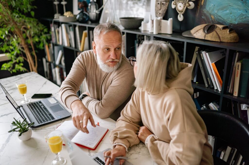 An older couple discussing paperwork together at a table with a laptop and documents.