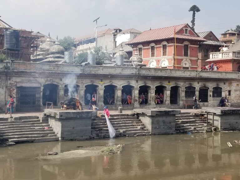 A riverside cremation site with stone steps, traditional buildings, and smoke rising near the water.