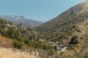 A mountain valley with dry golden grasses, green shrubs, and rocky slopes under a clear blue sky.