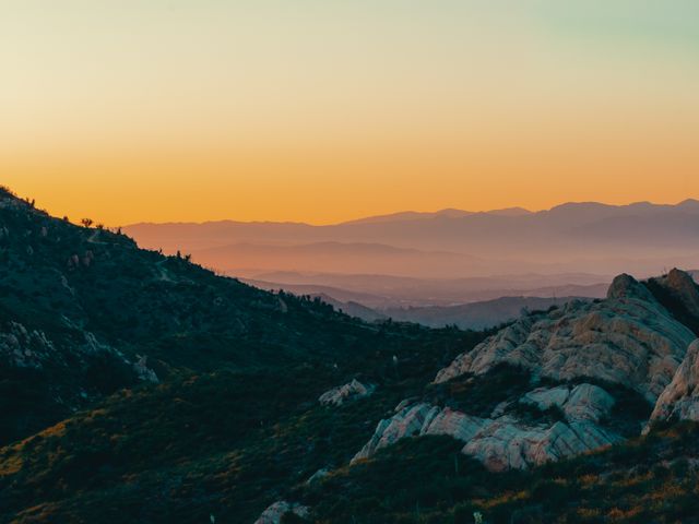 A rugged mountain landscape beneath a warm orange sunset, with distant ridges fading into the horizon.