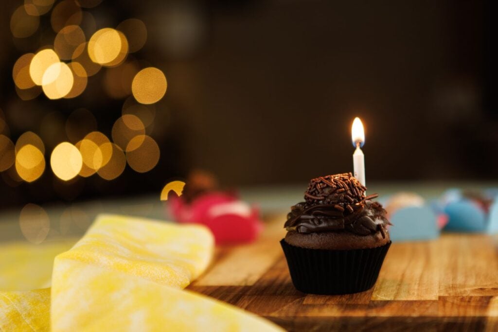 A chocolate cupcake with a lit candle sits on a wooden table, surrounded by soft, warm lights in the background.