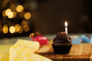 A chocolate cupcake with a lit candle sits on a wooden table, surrounded by soft, warm lights in the background.