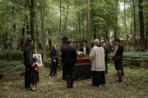 A small group of mourners gathers around a wooden coffin during an outdoor funeral service in a peaceful woodland setting.