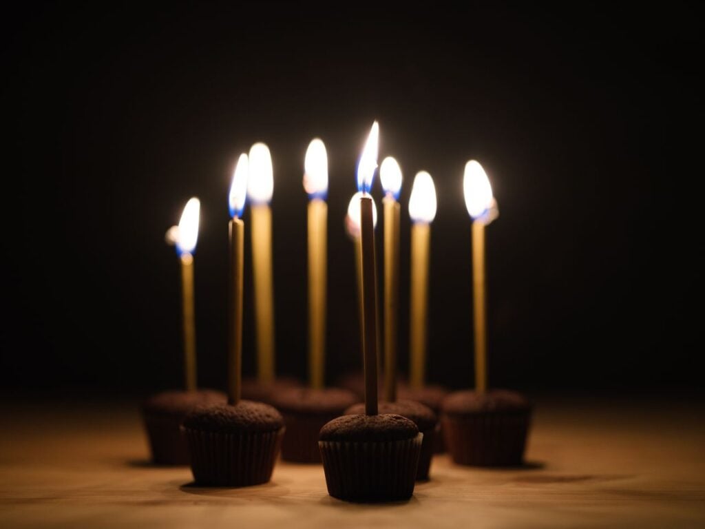 Several lit candles and chocolate cupcakes rest on a wooden table against a dark background.