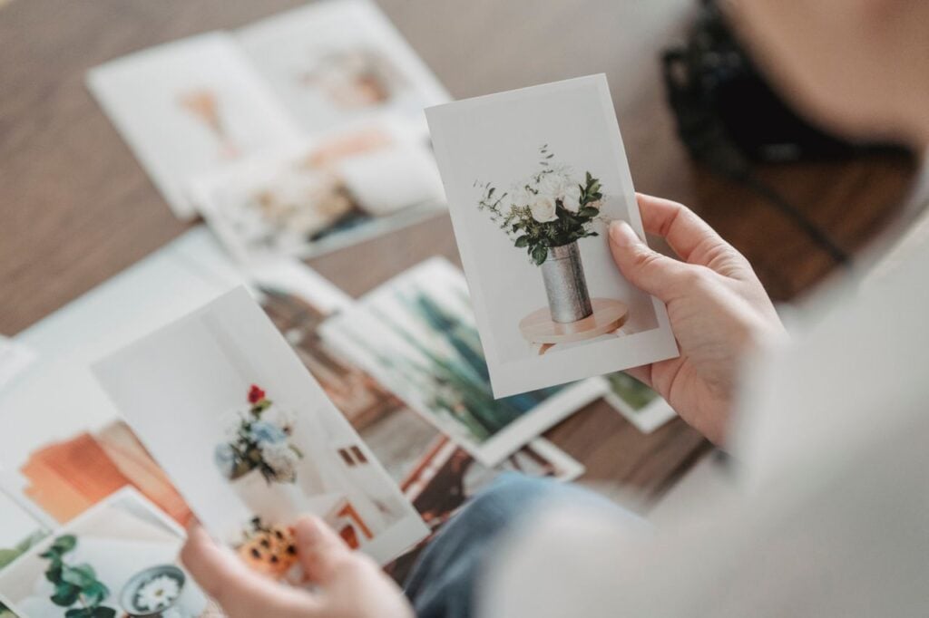A person looks through printed photographs, holding a picture of white flowers in a vase.