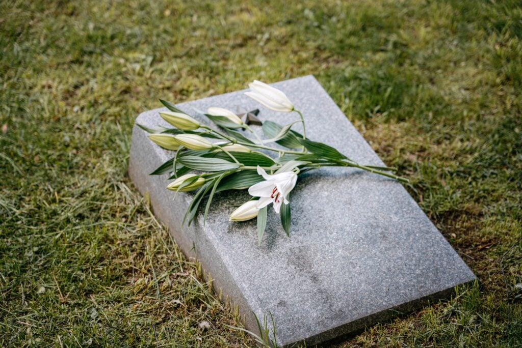 White lilies and greenery rest on a gray gravestone surrounded by grass.
