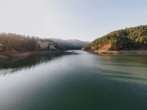 A peaceful lake surrounded by forested hills, with calm waters reflecting the soft light of the sky.