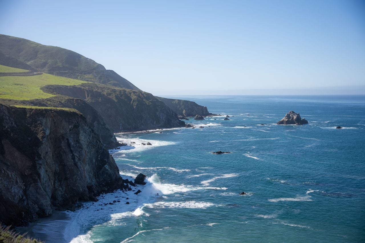 Rocky coastal cliffs overlooking the blue ocean, with waves breaking along the shoreline under a clear sky.