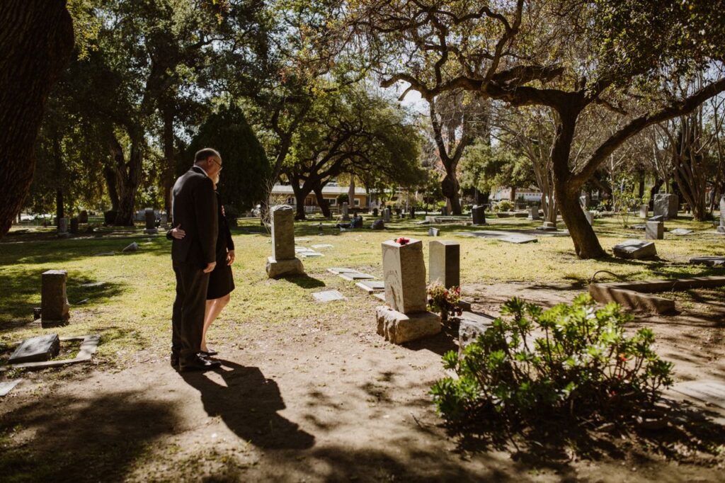 A couple stands quietly beside a grave in a peaceful cemetery surrounded by trees and sunlight.