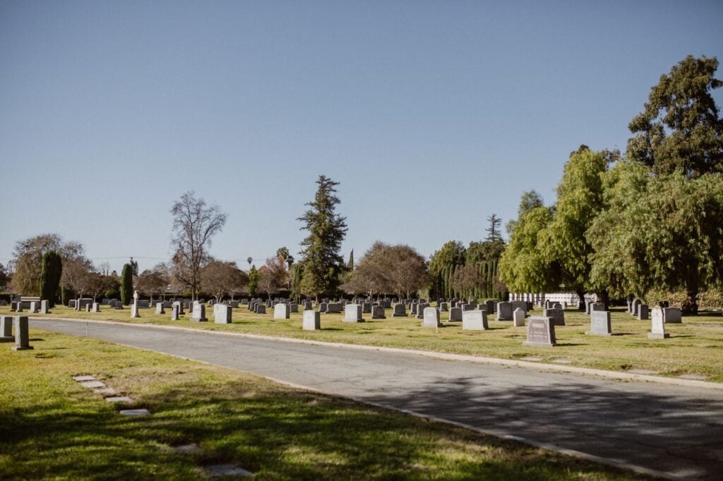 A quiet cemetery with rows of gravestones, green lawns, and tall trees beneath a clear blue sky.