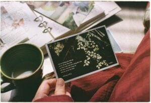 A person holds a floral postcard beside a green mug and an open scrapbook.