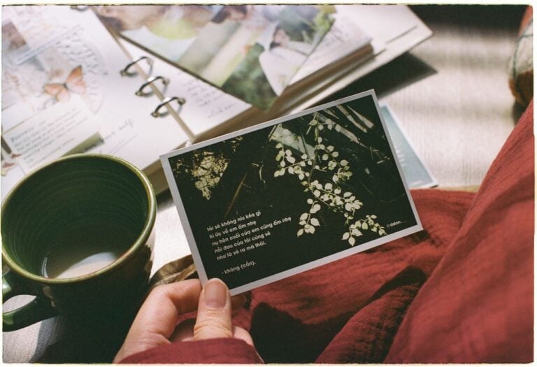 A person holds a floral postcard beside a green mug and an open scrapbook.